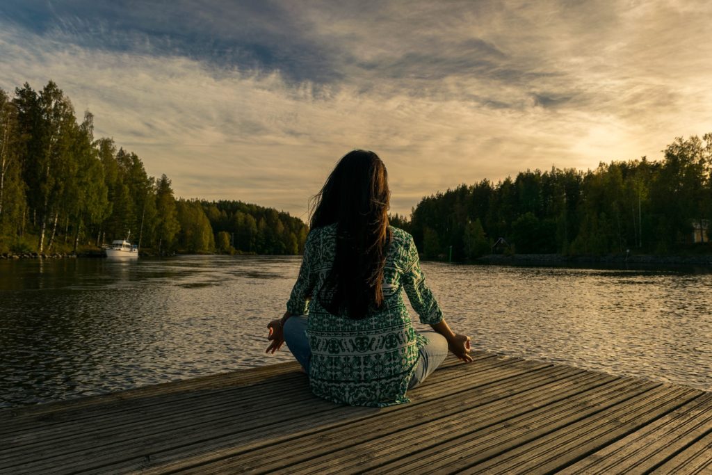 nervous system stuck in fight or flight, image of Person sitting on a dock in a meditative pose facing mountains and a body of water that’s golden in the sunlight—a representation of meditation for restoring a nervous system stuck in fight or flight.