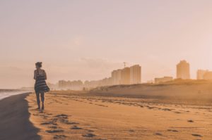 A women walking along a clear stretch of beach, with a cityscape in the background.