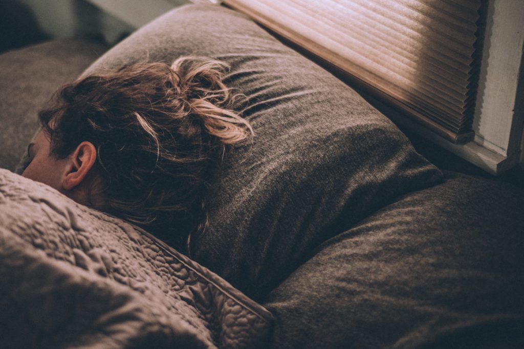 root cause of hair loss, Up-close photo of a person’s head resting on a pillow as they sleep, with long, thin, ashen blonde hair tied up in a messy bun—representing insufficient sleep as a potential root cause of hair loss.
