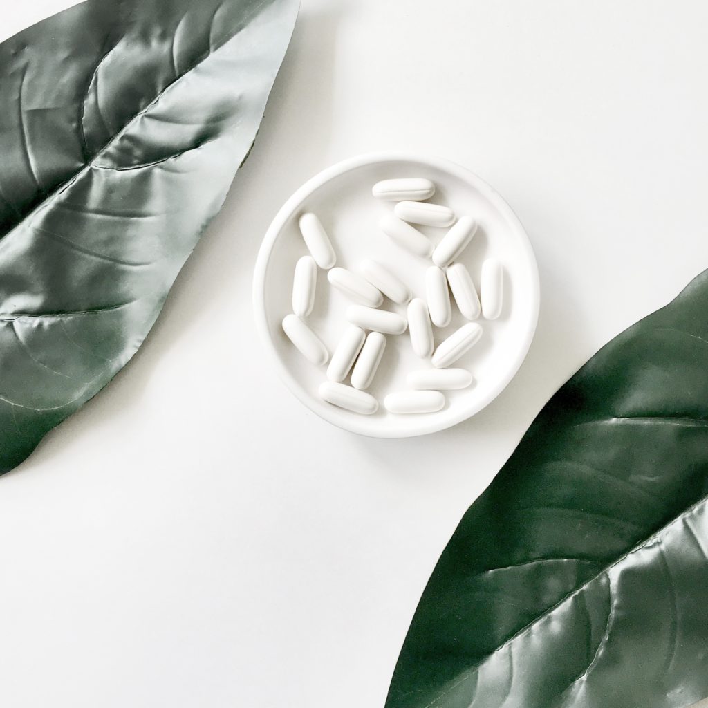 root cause of hair loss,Photo, shot from above, of a white plate filled with white tablet pills, sitting on a white table, with large, dark green leaves on either side of it—portraying natural supplementation as a possible way to address the root cause of hair loss.